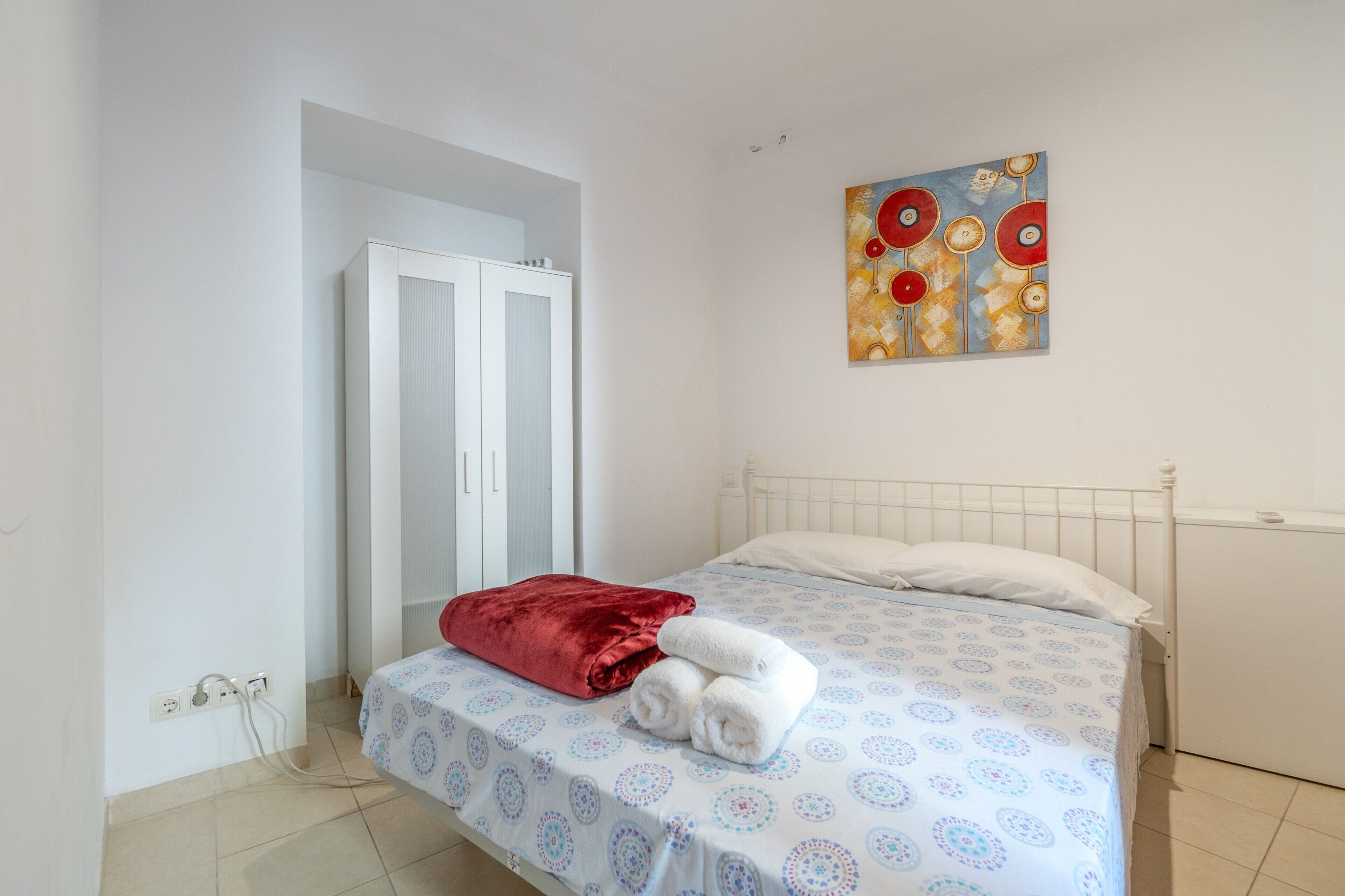 Un dormitorio con una cama, almohada roja, manta blanca, un cuadro en la pared y un armario blanco.