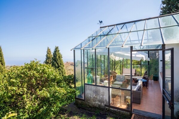 A greenhouse with a glass roof, a seating area with a table and chairs, and a view of the surrounding greenery.