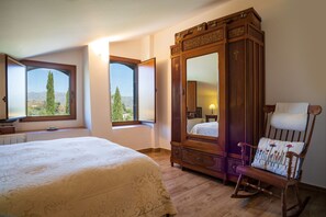 A bedroom with a large bed, a wooden wardrobe, a rocking chair, and a view of trees through a window.