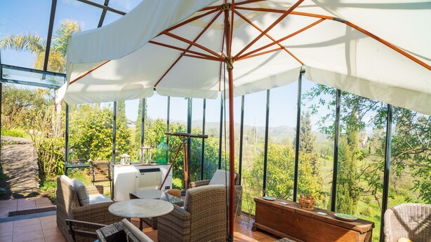 A covered outdoor seating area with wicker furniture, a wooden table, and a view of greenery. - Holiday Home 'Etnalodge' with Sea View, Private Terrace and Wi-Fi (Piedimonte Etneo)