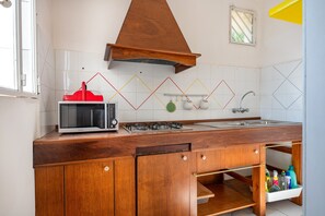 Een keuken met een magnetron, kookplaat, spoelbak, houten keukenkastjes en een raam.
