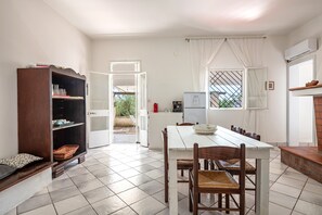 Een eetgedeelte met een witte tafel en stoelen, een houten kast en uitzicht door een open deur.