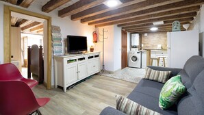 Un salón con sofá gris, sillón rojo y televisor sobre un mueble. Se observa una lavadora y una pequeña cocina al fondo.