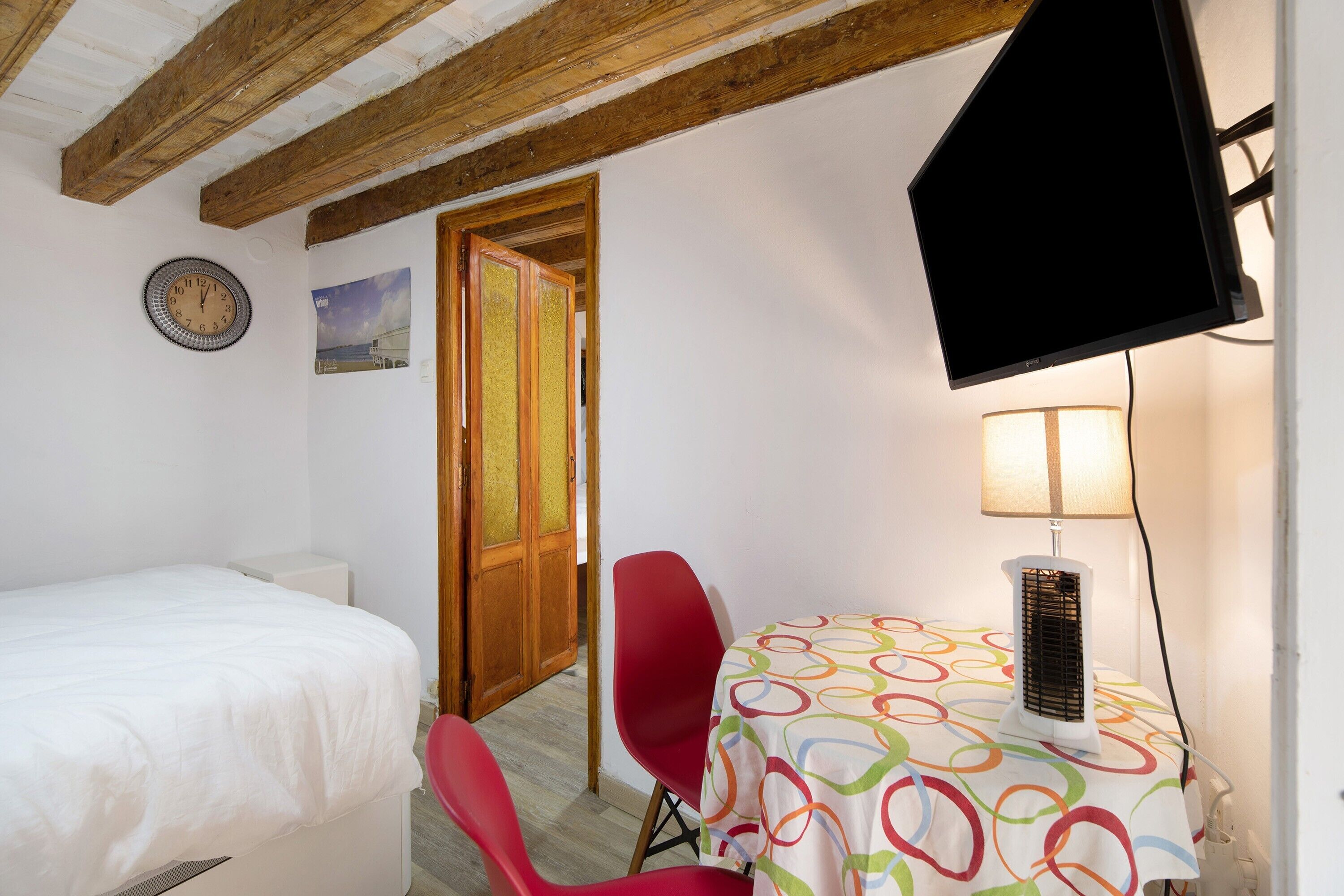Ein Zimmer mit einem Bett, einem roten Sessel, einem Tisch mit bunten Tischdecken, einer Wanduhr und einem Fernseher.