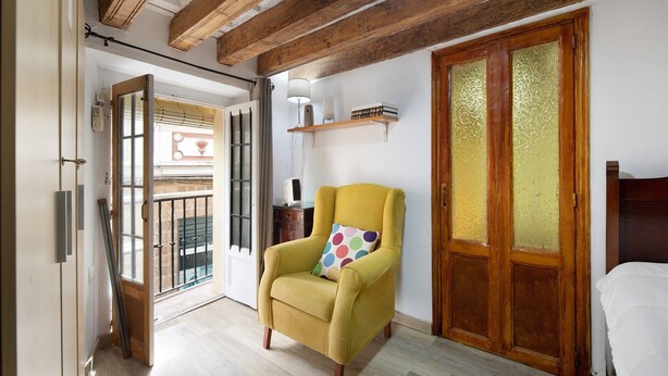 Una habitación con un sillón amarillo, vigas de madera en el techo, una puerta de madera con paneles de vidrio y una cama con ropa blanca.