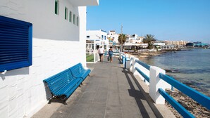 A coastal walkway with benches, a white building with blue shutters, and a clear view of the sea. - Vacation Apartment 'Central Bright Studio' (Corralejo)