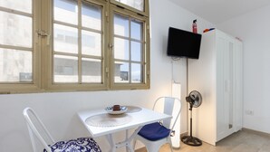 A room with a white table, two chairs, a standing fan, a TV on the wall, and a window with a view of buildings. - Vacation Apartment 'Central Bright Studio' (Corralejo)