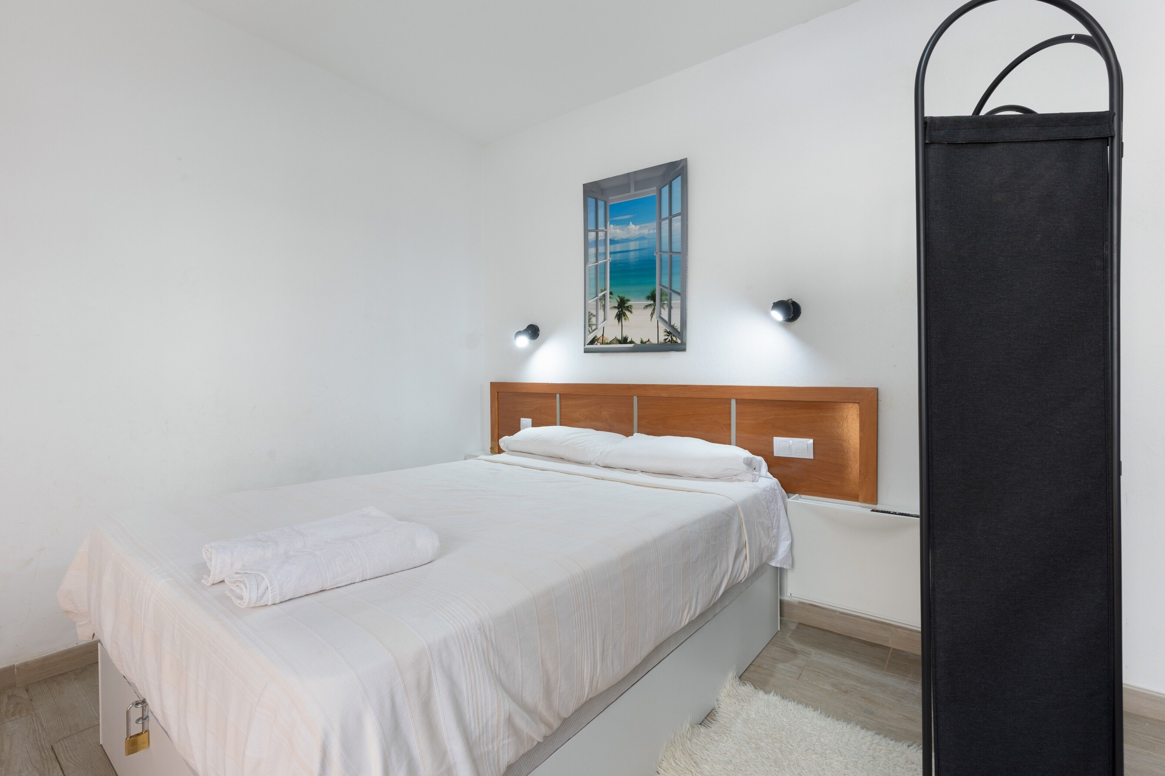 Un dormitorio con una cama, un cuadro de una playa, un cabecero de madera y un objeto negro en la pared.