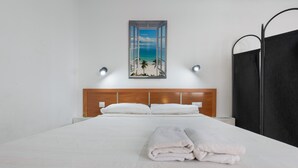 A neatly made bed with white linens and towels, a wooden headboard, a wall-mounted painting of a beach, and a black partition. - Vacation Apartment 'Central Bright Studio' (Corralejo)