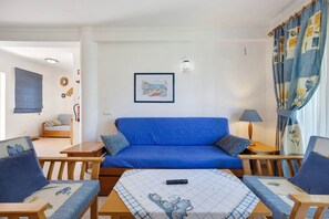 A room with a blue sofa, two wooden chairs, a small table with a remote control, a lamp, and a picture on the wall.