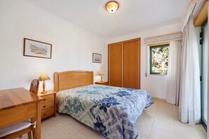 A bedroom with a bed, wooden nightstands, a desk, and chairs, a window with curtains, and two framed pictures on the wall.