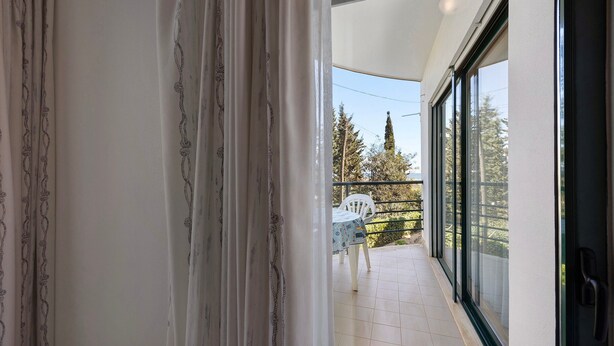 A balcony with a table and chairs, a view of trees, and a door leading to the interior.