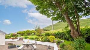 A patio area with a picnic table, surrounded by lush greenery and a hillside in the background.