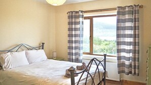 A bedroom with a bed, a window with curtains, a radiator, and a view of greenery outside.