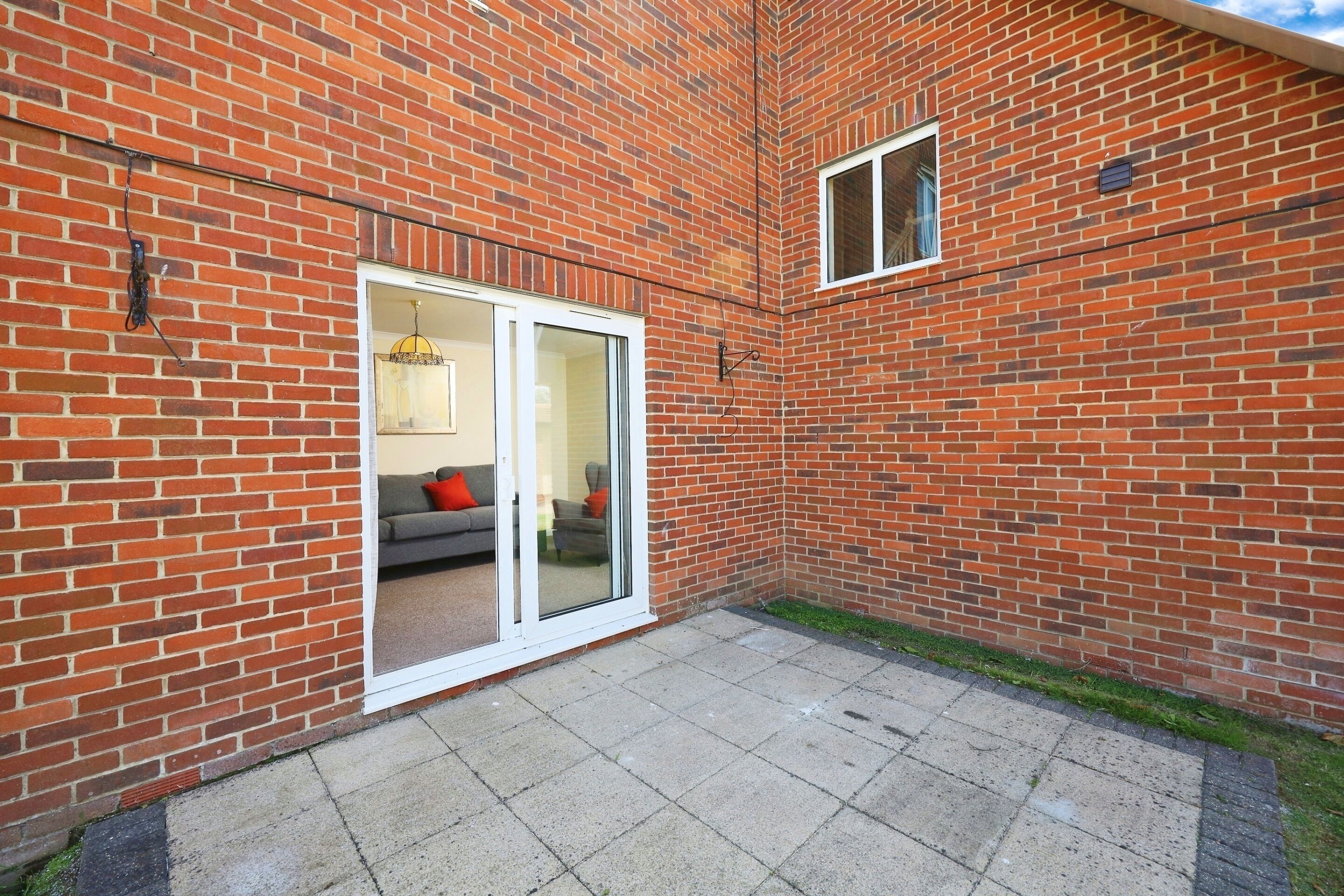 A brick building with a patio area, a sliding glass door leading to an interior space with a sofa and cushions, and a small window on the upper right.