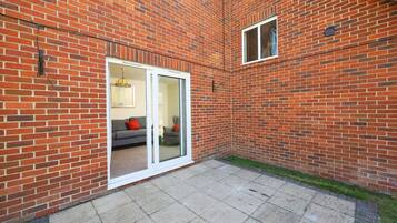 A brick building with a patio area, a sliding glass door leading to an interior space with a sofa and cushions, and a small window on the upper right.