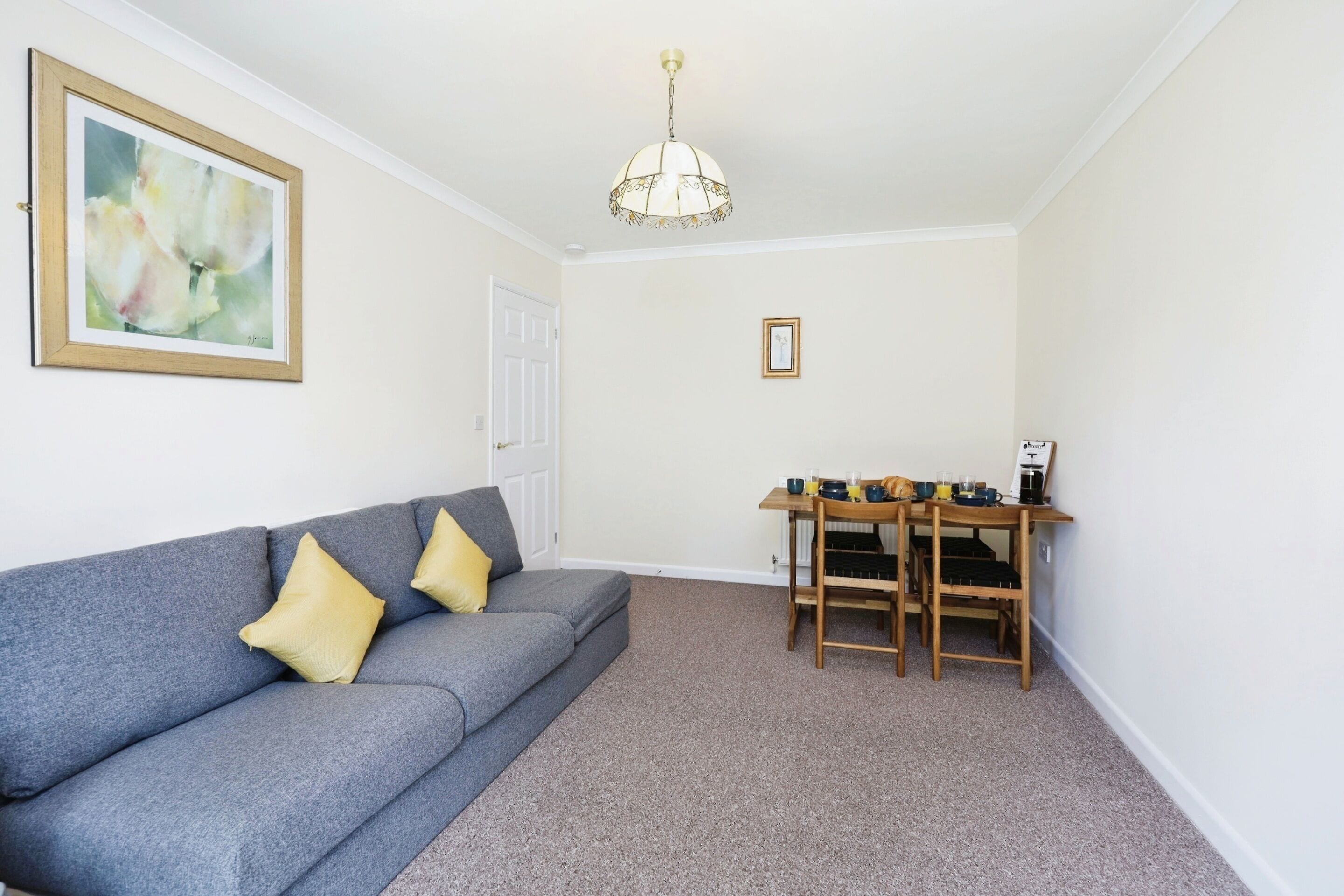 A living room with a grey sofa, two yellow cushions, a wooden table with chairs, a coffee machine, and a framed picture on the wall.