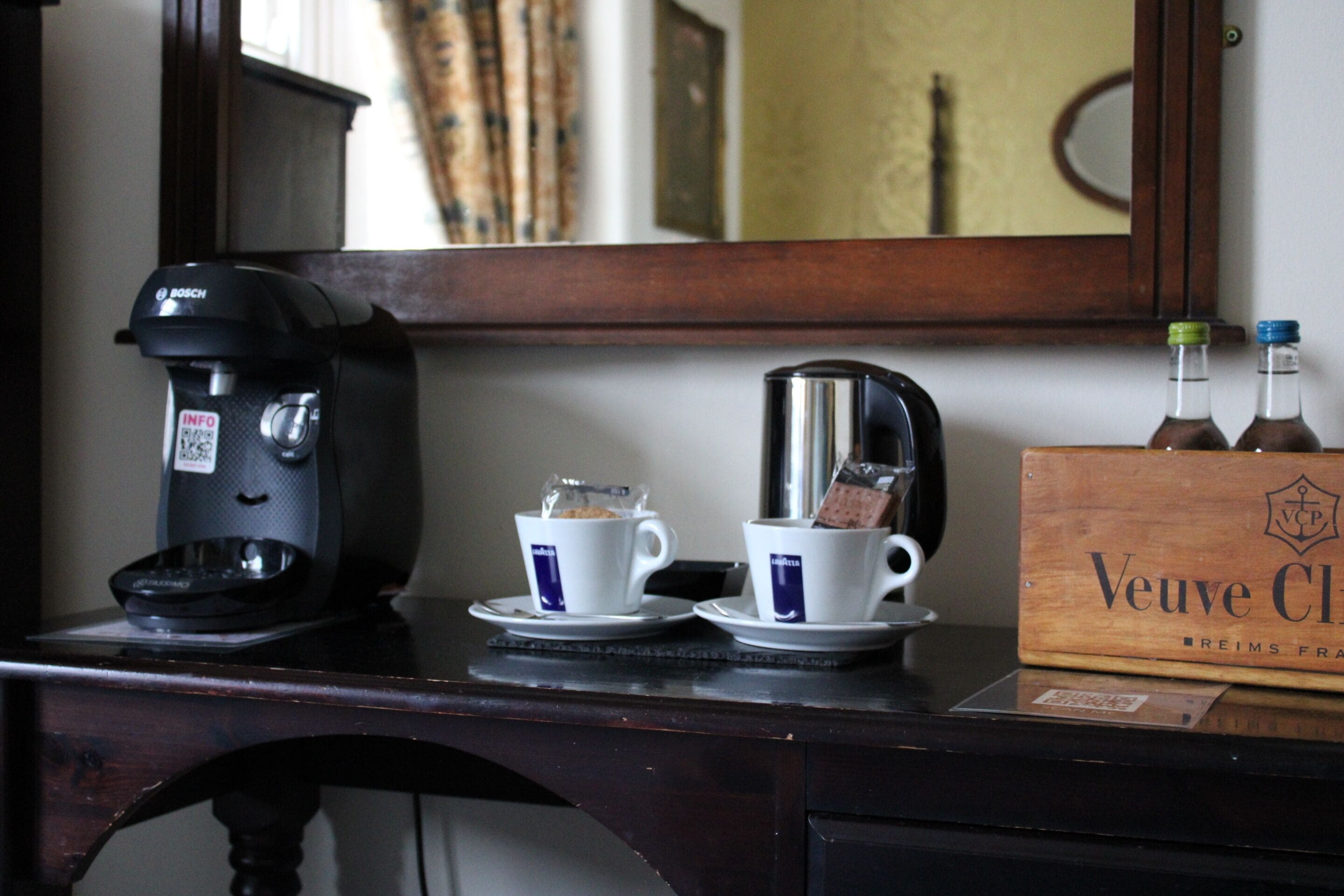 A coffee maker, two cups with tea bags, and a box labeled "Veuve Clicquot" on a wooden table.