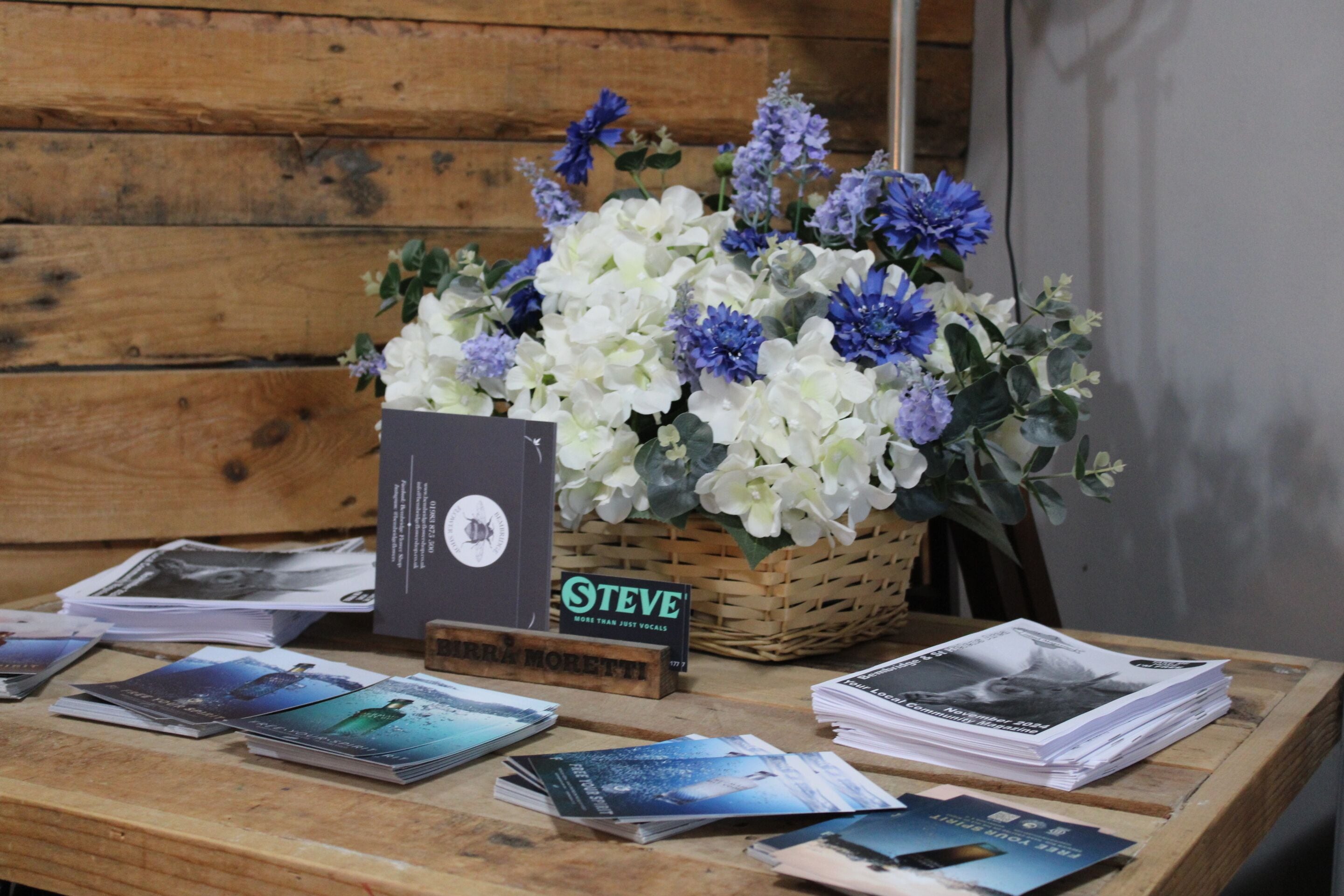 A wooden table with a basket of flowers, a sign, and some brochures.