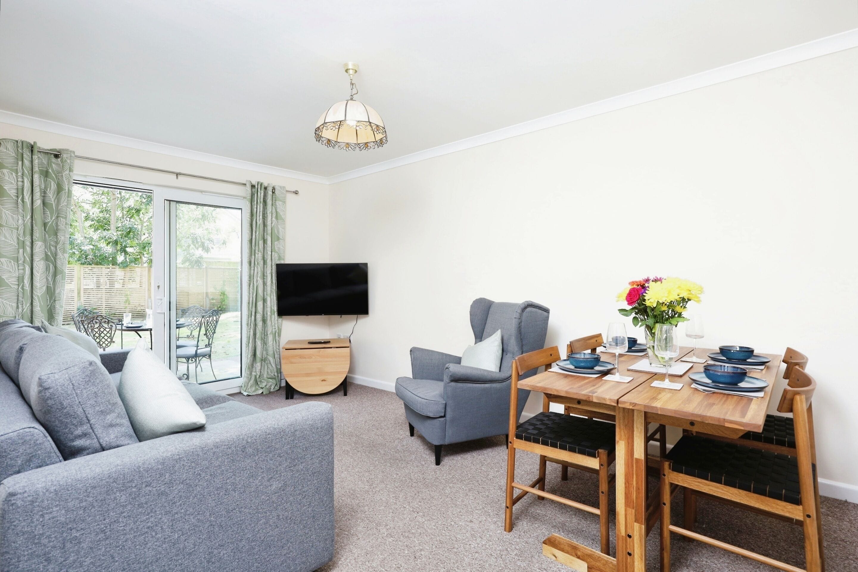 A modern living room with a grey sofa, a dining table set with blue bowls and a vase of flowers, and a flat-screen TV on the wall.
