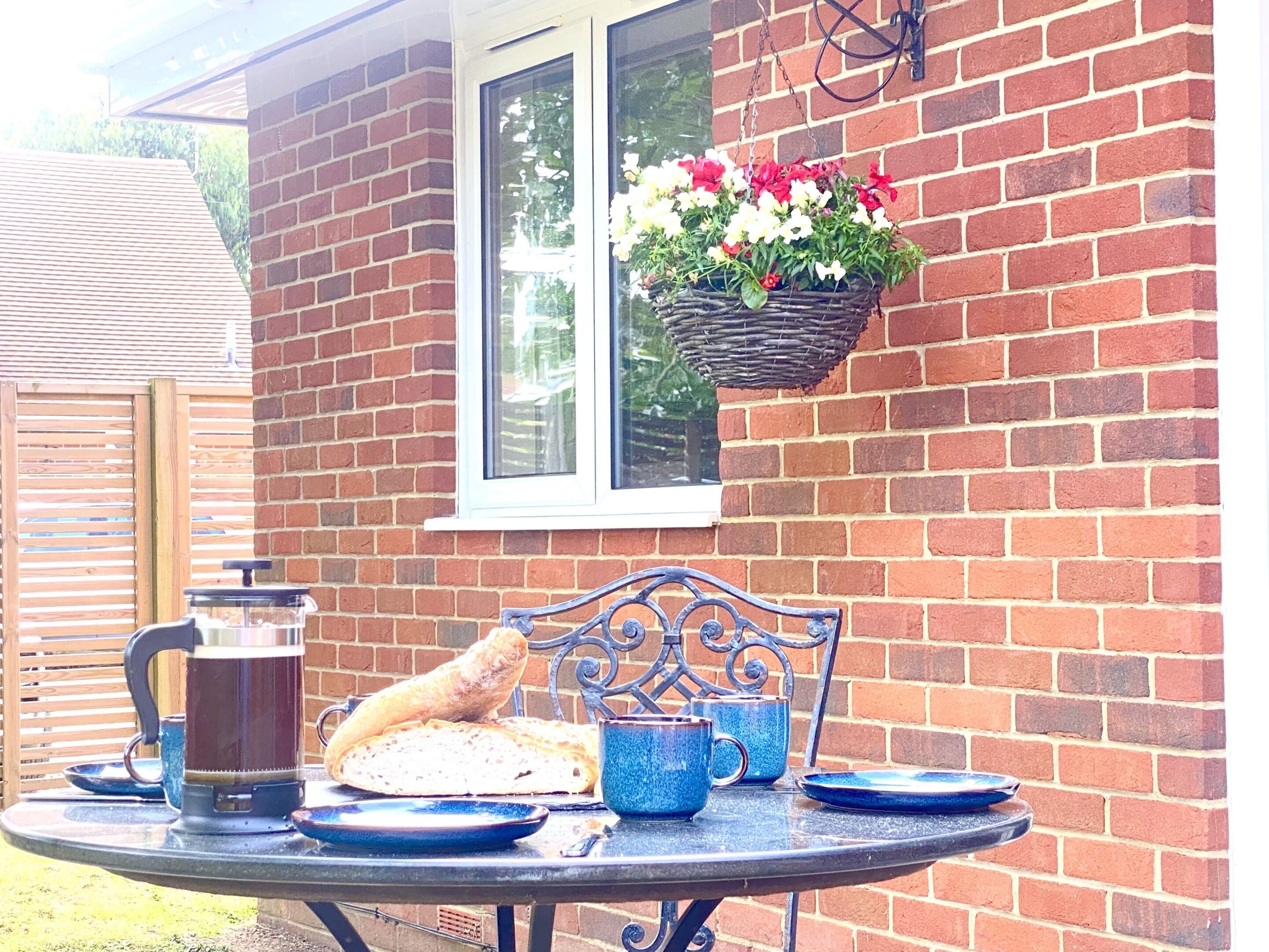 A table set with bread, a coffee pot, and blue cups, with a hanging flower basket on a brick wall.