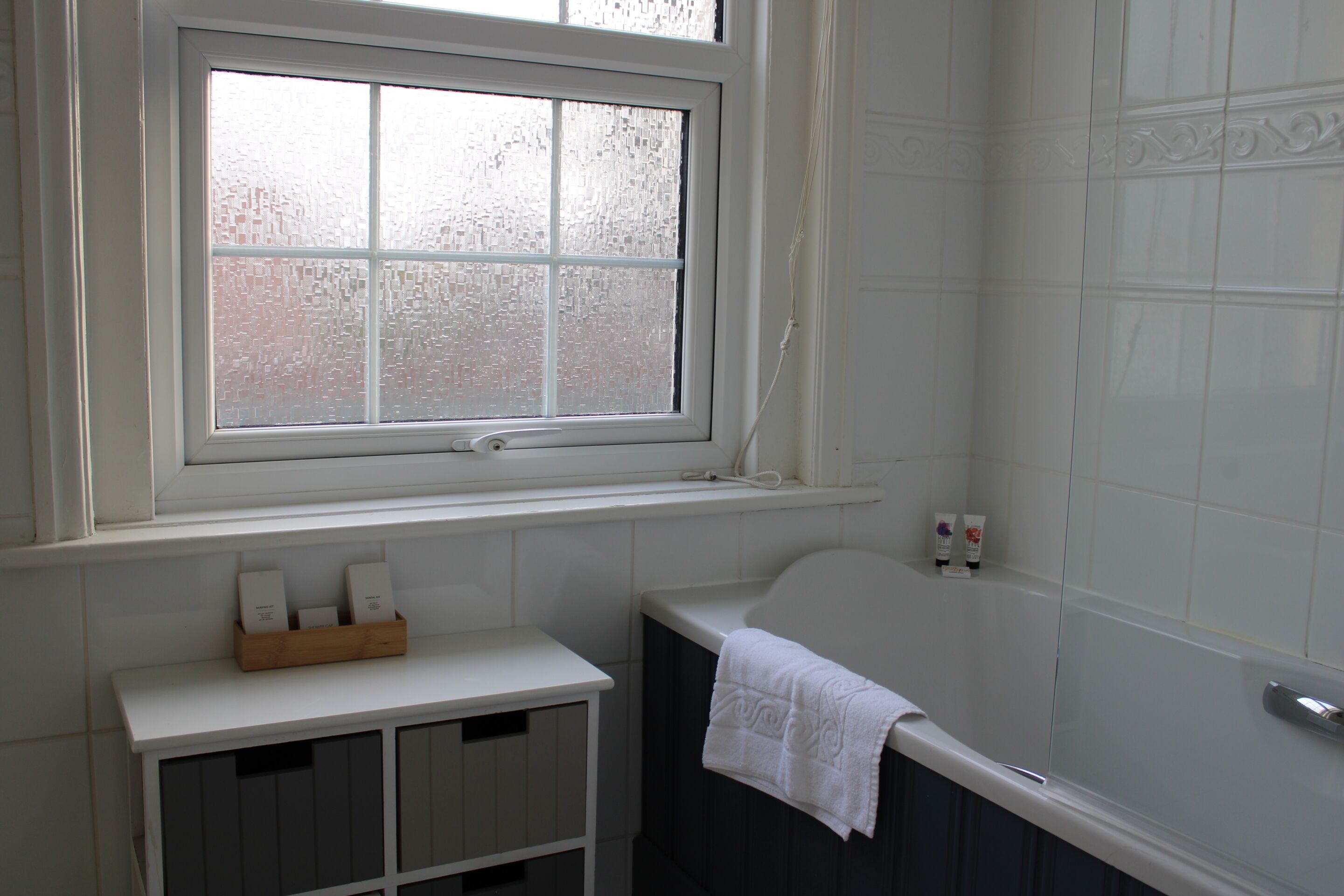 Une salle de bain avec une fenêtre, une baignoire et une petite étagère où sont rangés les articles de toilette.