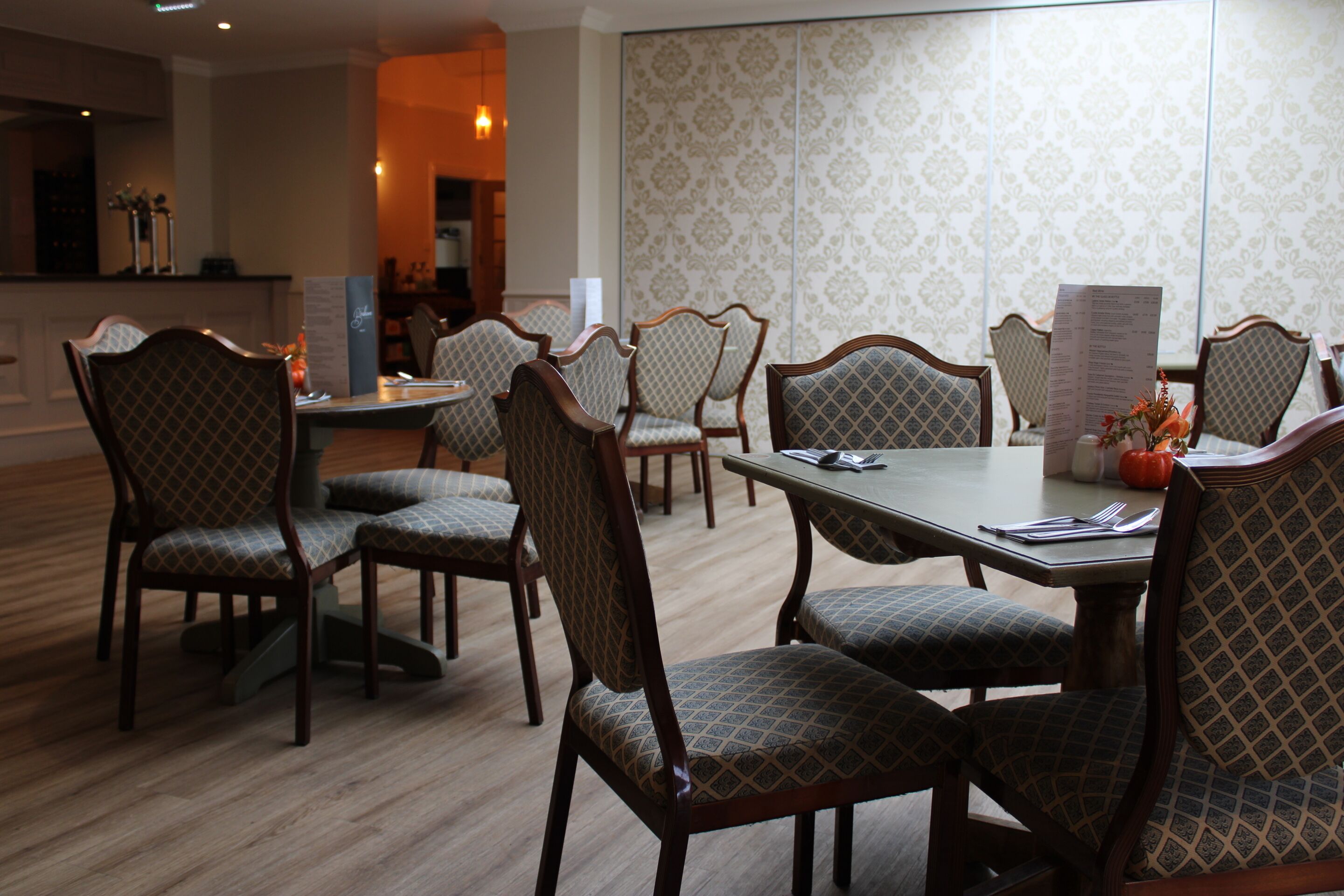 A dining area with wooden chairs and tables, a menu stand, and a patterned wallpaper wall.