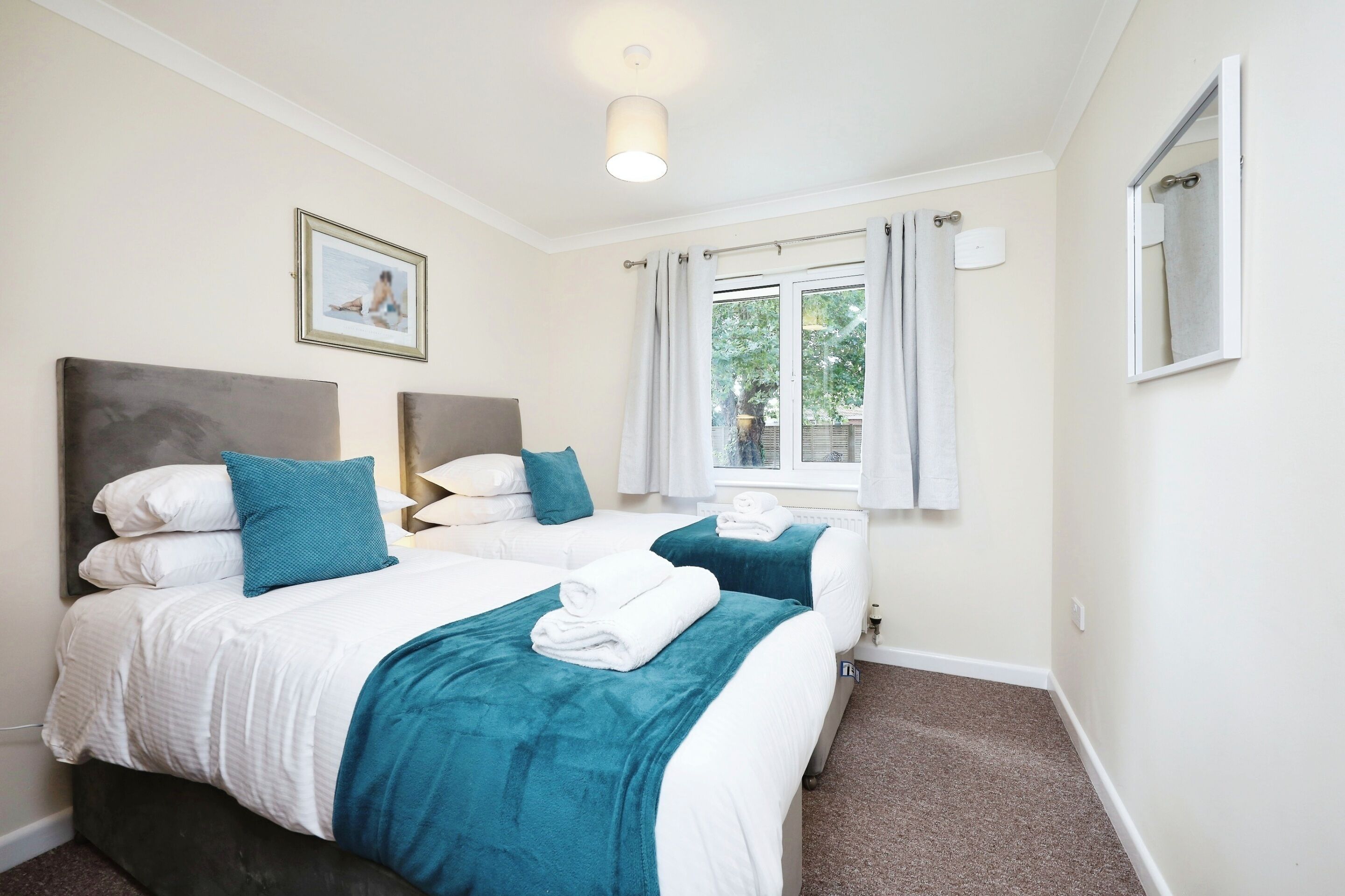 A hotel room with two beds, each with white bedding and teal throw pillows. There is a window with curtains, a ceiling light, and a framed picture on the wall.