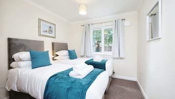 A hotel room with two beds, each with white bedding and teal throw pillows. There is a window with curtains, a ceiling light, and a framed picture on the wall.