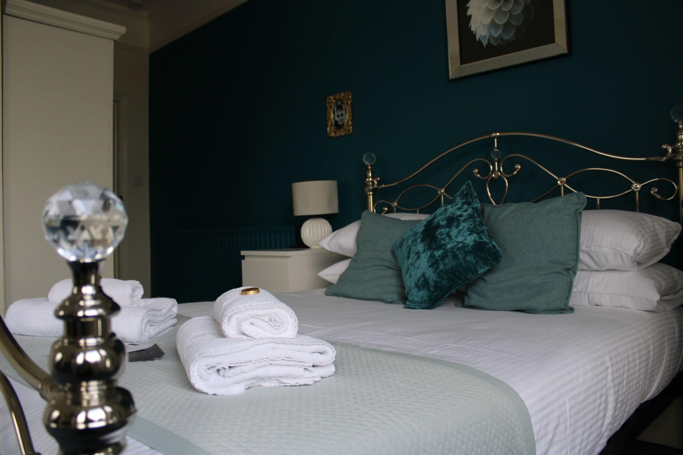 A neatly made bed with white linens and teal pillows, a bedside lamp, and a framed picture on the wall.