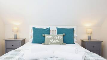 A neatly made bed with a pillow that reads "our happy place," two bedside lamps, and a simple gray nightstand.