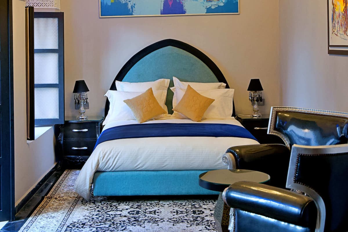 A bedroom with a large bed, a chandelier, a colorful abstract painting, a patterned rug, and a leather armchair.