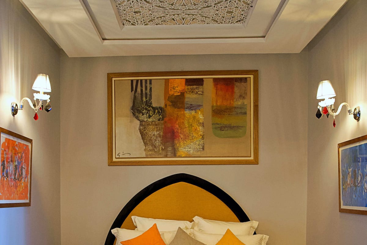 A neatly made bed with a patterned bedspread and pillows, a large abstract painting above it, and a detailed carpet.