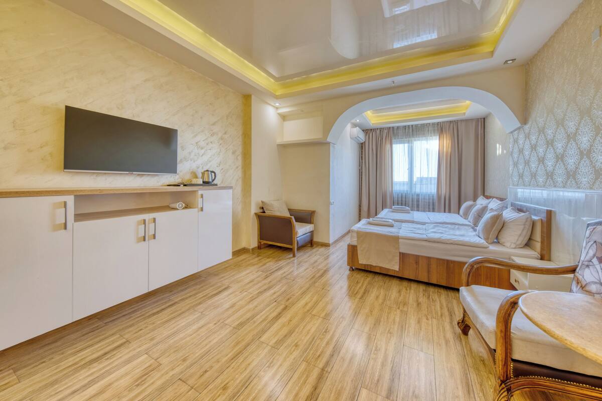 A modern hotel room with a large bed, a TV on the wall, a wooden floor, and a small seating area.