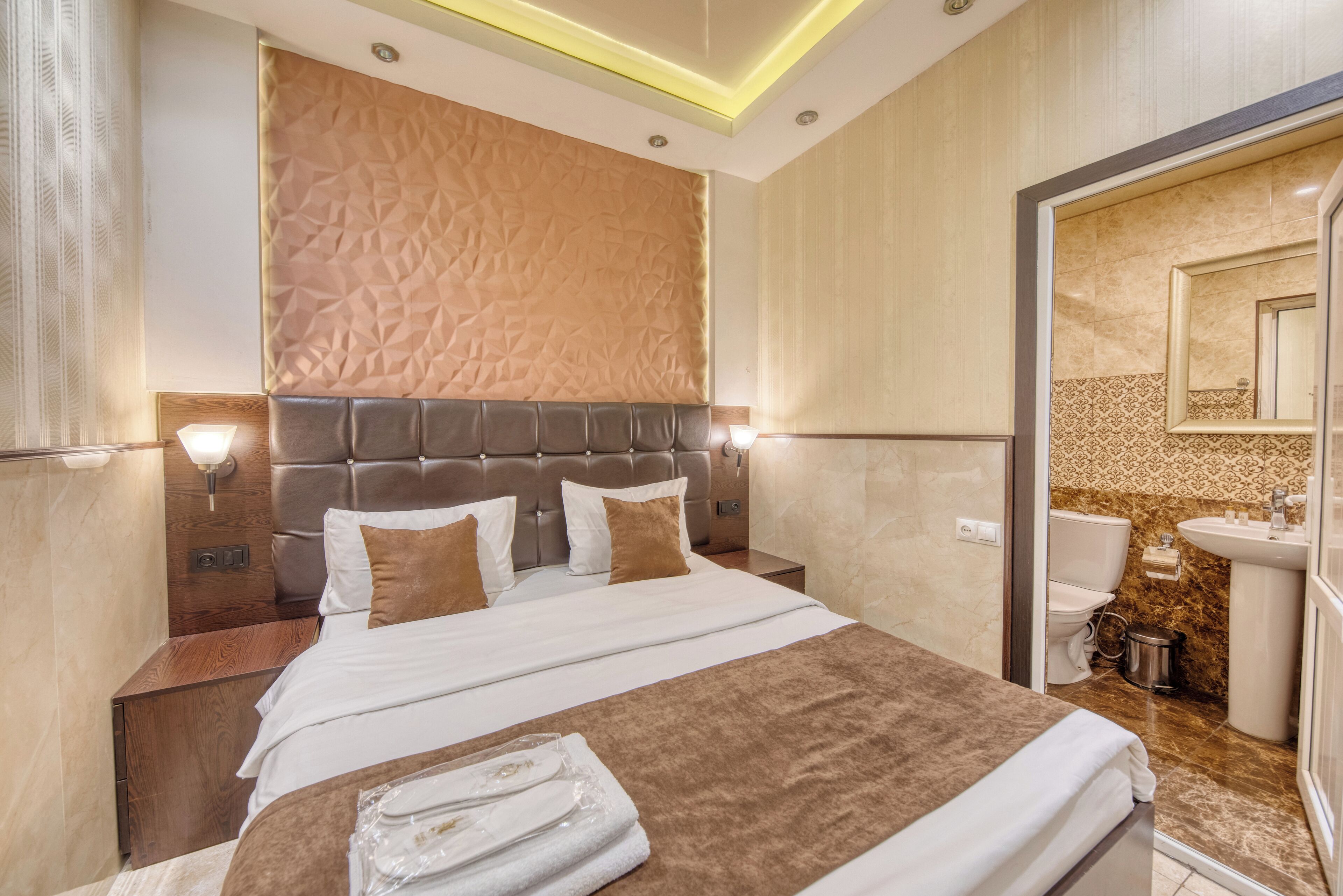 A modern hotel room with a large bed, a headboard with a geometric pattern, a bathroom with a marble countertop, and a mirror above the sink.