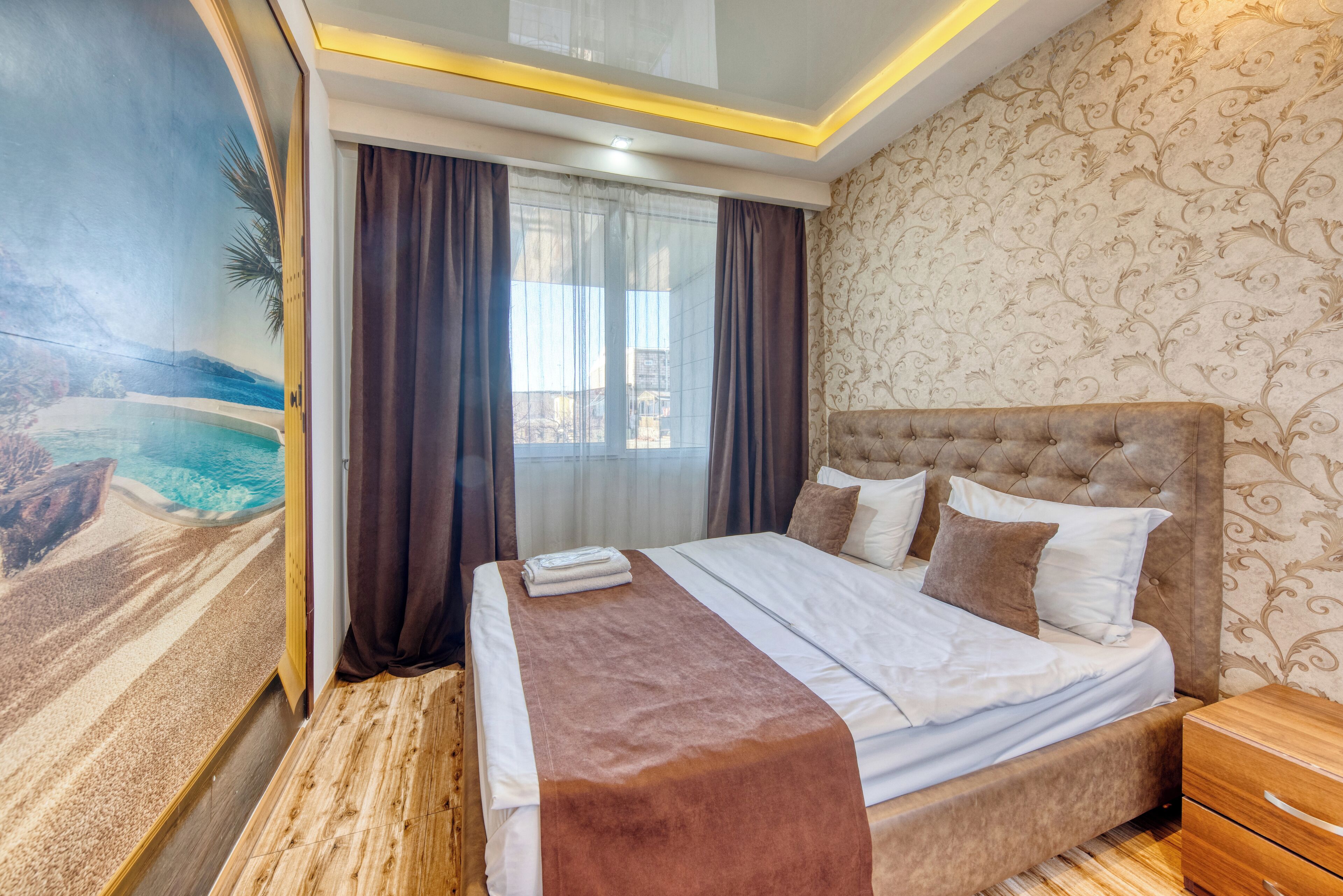 A hotel room with a large bed, a wooden floor, a view of a mountainous landscape through a window, and a patterned wallpaper on the wall.