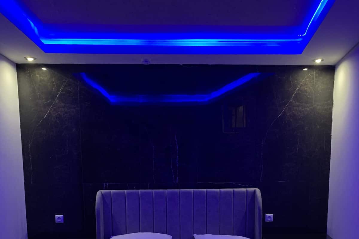 A modern bedroom with a large bed, dark walls, and blue ambient lighting.