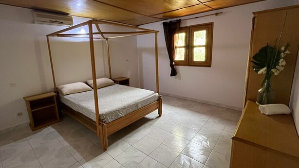 Un dormitori amb un llit amb cortines, una petita taula de nit, una meseta de nit, una finestra amb cortines i una planta sobre una petita estant.