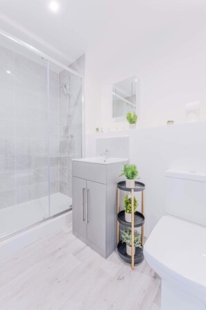 A modern bathroom with a glass shower enclosure, a grey cabinet with a sink, and a black shelf with plants. - Smart 1 Bed Apartment in Wolverhampton (Wolverhampton)