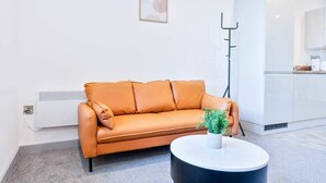 Un salon moderne avec un canapé, une petite table ronde agrémentée d’une plante, un lampadaire et, au fond, un espace cuisine blanc.