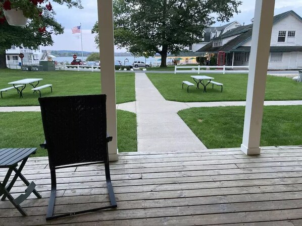 A covered patio with picnic tables and a black chair, overlooking a grassy area with a pathway leading to a dock.