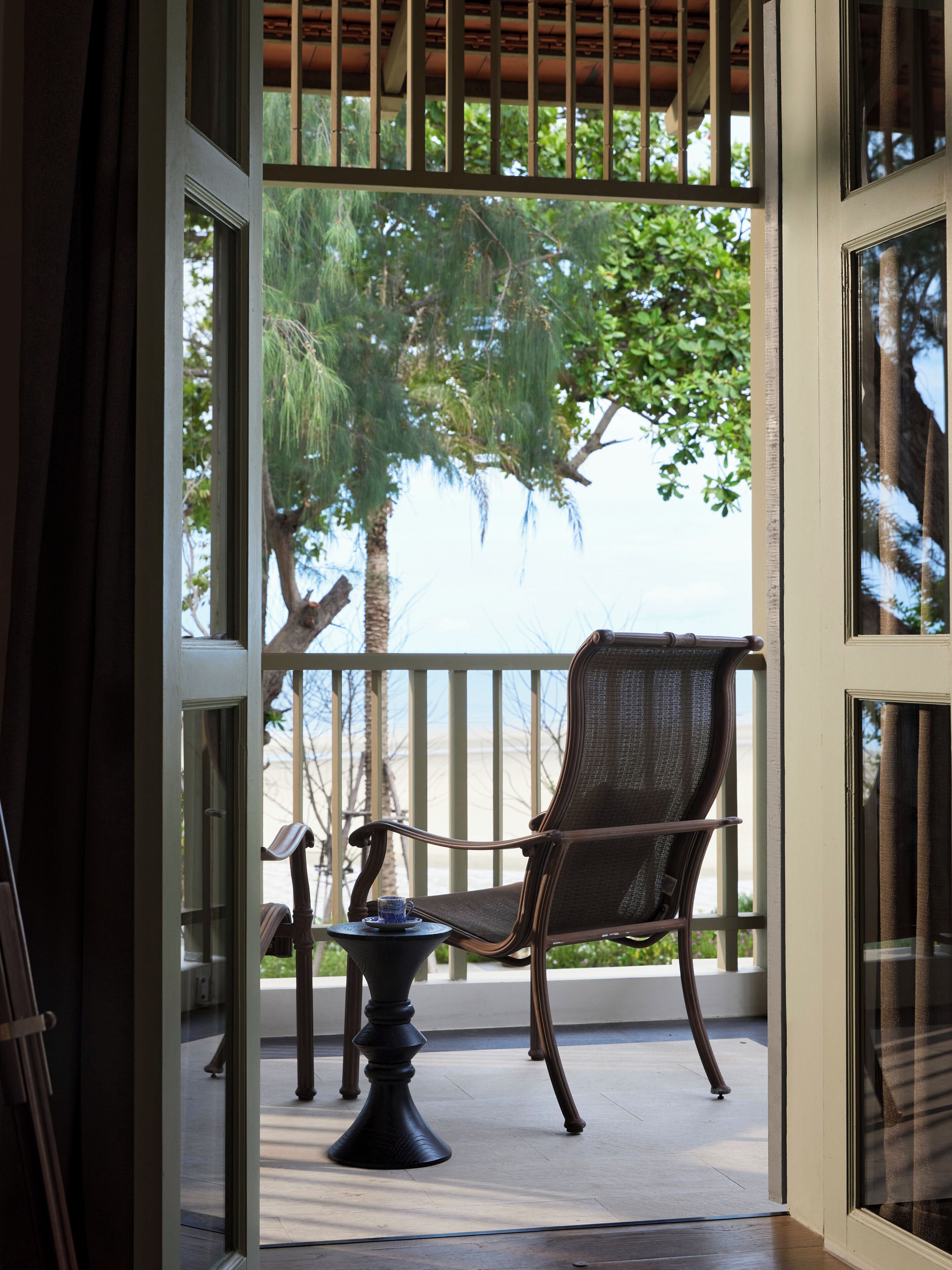 A balcony with a wicker chair and a small table, overlooking a palm tree and a beach.