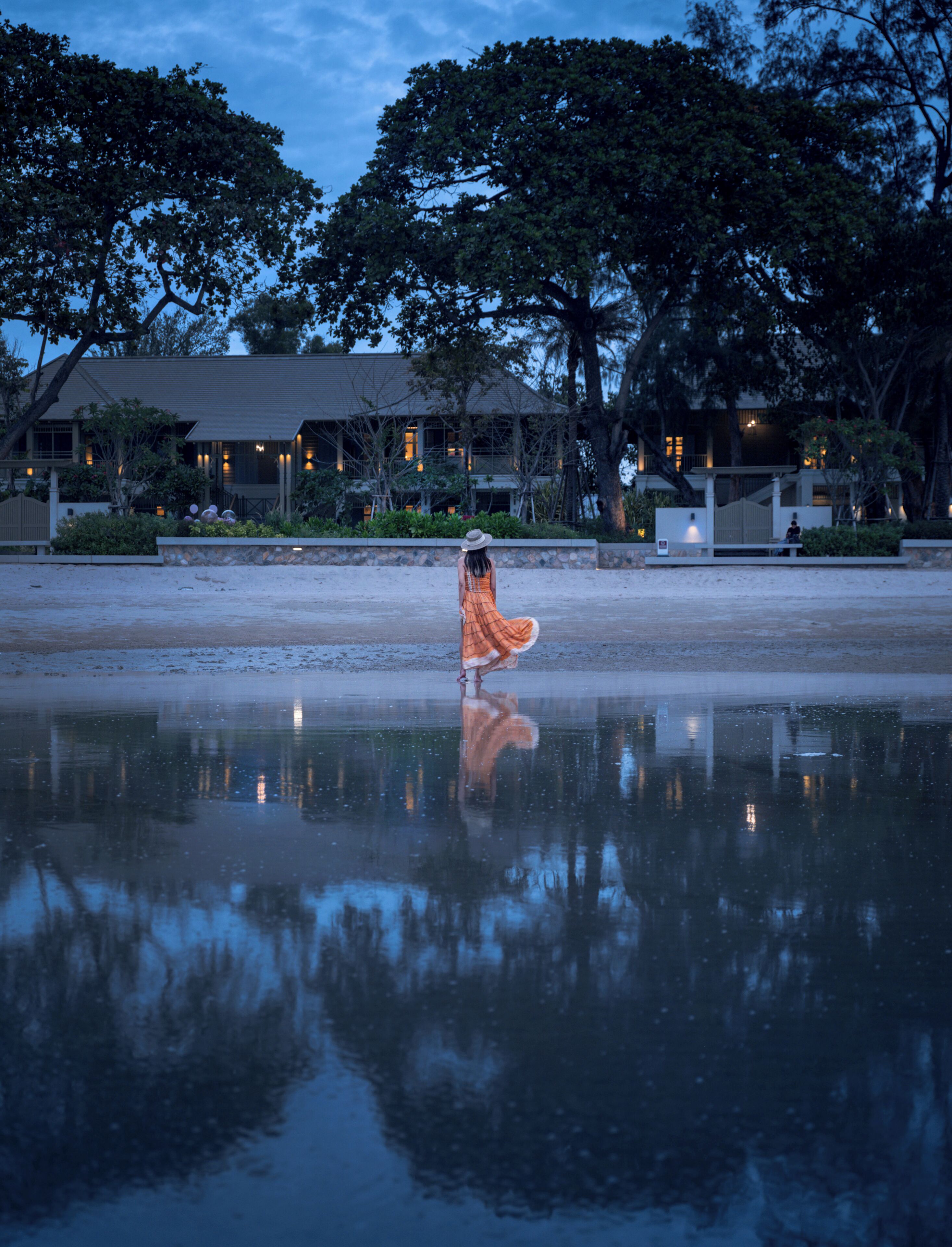 A reflective water surface with a person in an orange dress and a hat, a building with multiple windows, and trees in the background.