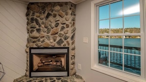 A stone fireplace with a log fire, a window with a view of a dock and water, and a stone wall.