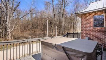 A deck with a hot tub, a brick building, and a forested area in the background.