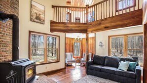 A cozy living room with a fireplace, wooden floors, a sofa, and large windows. - Private 40-Acre Retreat | Hot Tub, Trails, Games & Fire Pit (Hopkins)