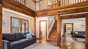 A spacious living area with a dark gray sofa, patterned rug, wooden flooring, and a staircase leading to an upper level. - Private 40-Acre Retreat | Hot Tub, Trails, Games & Fire Pit (Hopkins)