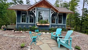 Una cabaña con un porche delantero, un patio de piedra, dos sillas Adirondack y una jardinera con flores.