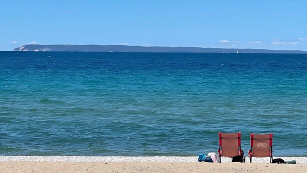 Una playa con dos sillas rojas y una bolsa sobre la arena, frente a un mar tranquilo y una tierra distante.