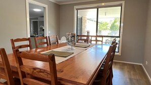 A dining room with a wooden table and chairs, a glass vase with flowers, and a view of the outdoors through a large window. - Sandy Retreat on Royal Inverloch (Inverloch)